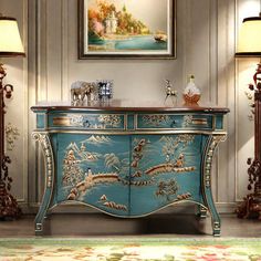 a blue and gold painted dresser with two lamps on it's sides, in front of a painting