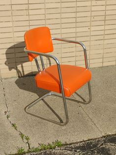 an orange chair sitting on the sidewalk next to a brick wall with grass growing in it