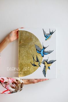 two hands are holding up a painting with birds on it and the bottom half is gold