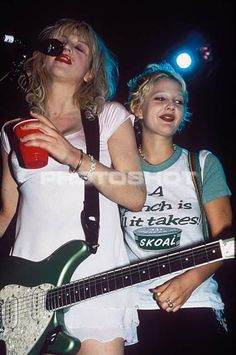 two people standing next to each other while one holds a guitar and the other is holding a cup