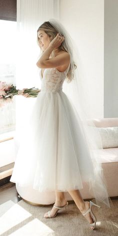 a woman in a white wedding dress is standing near a window and holding her hand up to her face