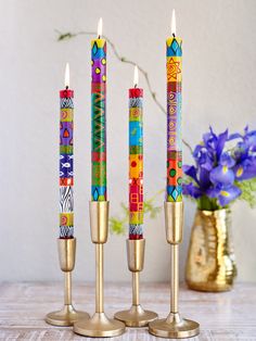 three candles with colorful designs on them sitting in front of a vase and purple flowers