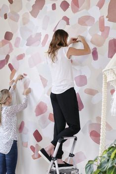 two women are painting a wall with pink and white shapes on it, while one woman is standing on a ladder