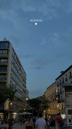 two people walking down the street in front of some tall buildings at night with lights on