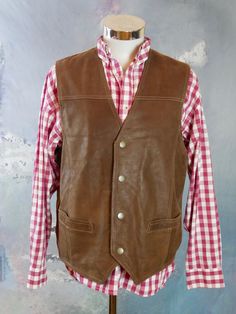 This 1990s brown leather vest has a V neckline, and closes in the front with four snap buttons. The vintage pointed-front Western waistcoat has two pockets on the front, and features topstitching on the leather. The vest has a full beige canvas (cotton) back with a leather half-belt with buckle. Chest = Up to 48 inches (121.92cm) Vest Length (front) = 26 inches (66.04cm) Vest Length (back) = 24 inches (60.96cm) Brand label: Pionier Sportive Size: 46 to 48 US/UK Material: Genuine Leather --- COND Classic Fall Vest With Snap Buttons, Fitted Brown Vest With Button Closure, Classic Leather Vest With Button Closure, Leather Vest With Button Closure, Vintage Leather Vest Outerwear, Fitted Vintage Leather Vest, Vintage Fitted Leather Vest, Leather Sleeveless Vest With Button Closure, Retro Brown Vest For Workwear