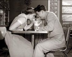 two people sitting at a table with cups in their hands and one person touching the other's face