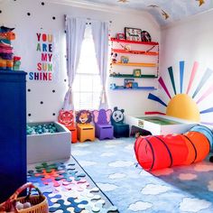 Bright blur rug with white fluffy clouds in center of room. Rainbow color shelves on a wall. Large horizon rising sun with rainbow colored beams. Large ball pit filled with an array of blue hued balls. Black and white polka dots on wall Playroom Ideas Colors, Playroom Ideas Rainbow, Bright Color Playroom, Playroom Bright Colors, Playroom Decor Colorful, Bright Rainbow Bedroom, Fun Colorful Nursery Ideas, Playroom Inspiration Colorful, Colorful Toy Room