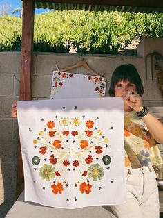 a woman sitting on a bench holding up a white piece of cloth with orange and yellow flowers