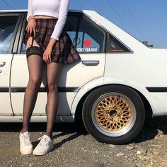 a woman standing next to a white car on the side of the road with her legs crossed