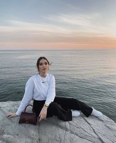 a woman sitting on top of a rock next to the ocean with a purse in her hand