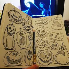 a hand holding an open sketch book with halloween faces on it and a tv in the background