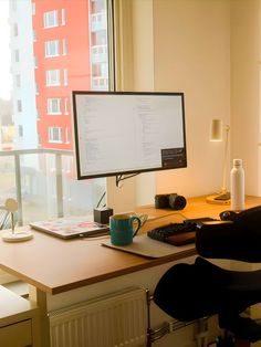 A minimalist home office setup featuring a large desk with a monitor, keyboard, and personal items. The workspace is bathed in natural light from a large window overlooking urban apartment buildings, creating a bright and inviting atmosphere. Minimal Aesthetic Home, Small Modern Apartment, Minimal Desk Setup, Male Living Space, Aesthetic Home Office, Modern Apartment Interior, Workspace Studio, Dream Desk, Computer Desk Setup