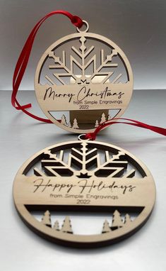 two wooden christmas ornaments with red ribbon on white table top, one has a snowflake ornament and the other says happy holidays