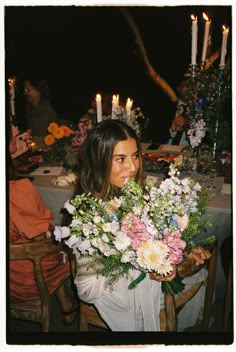 a woman sitting at a table with flowers in front of her and candles on the table behind her