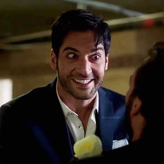 a man in a suit smiling and holding a yellow flower to his face while another man looks at him