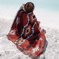 a person sitting on the beach with a blanket over their head looking out at the water