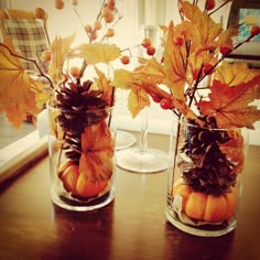 two glass vases filled with leaves and acorns on top of a wooden table