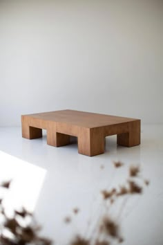 a square wooden table sitting on top of a white floor next to a vase with dried flowers