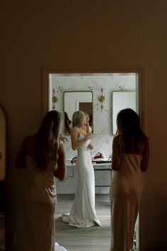 three women standing in front of a mirror looking at their wedding dresses and getting ready