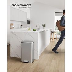 a man walking through a kitchen next to a white counter top with a silver trash can