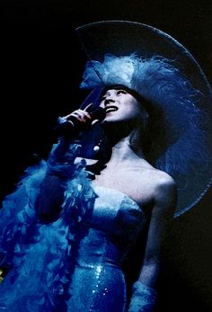 a woman in a blue dress and hat singing into a microphone with feathers on it