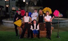 a group of people standing around each other in front of a fountain with umbrellas