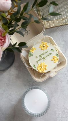 a white cake with yellow flowers on it sitting next to a candle and some pink roses