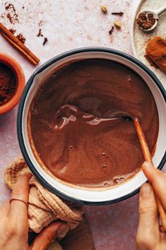 two hands holding a spoon over a bowl of chocolate pudding with cinnamons on the side