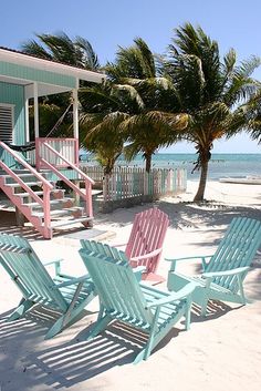 four lawn chairs sitting on the beach in front of a blue and pink house with palm trees