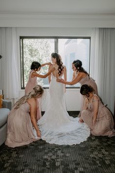 bridesmaids helping the bride put on her wedding dress in front of a window