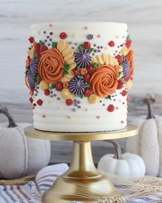 a white cake with orange and purple flowers on it sitting on a gold plate next to pumpkins