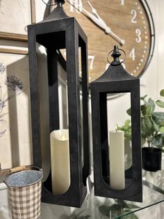 two black lanterns sitting on top of a glass table next to a clock and potted plant