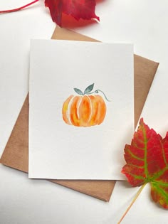 a card with a watercolor painting of an orange pumpkin on it next to some leaves