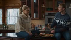 a man and woman sitting on a counter in a kitchen, one holding a pizza