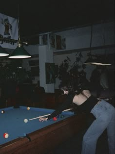 two people playing pool in a dark room with lights hanging from the ceiling and posters on the wall