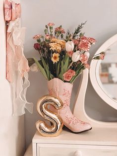 a vase filled with flowers next to a mirror and a gold s boot sitting on top of a white dresser