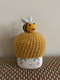 a crocheted cup with a smiling face and a bee on top, sitting on a table