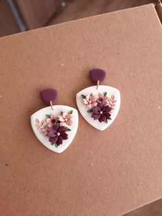 two white heart shaped earrings with purple flowers on them sitting on top of a cardboard box