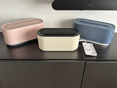 three different colored containers sitting on top of a black dresser next to a white wall