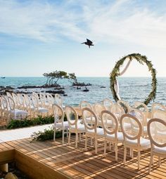 an outdoor ceremony set up with white chairs and greenery on the side of the water