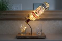 a lighted bottle lamp sitting on top of a wooden table next to two shot glasses