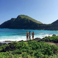 three people standing on the beach looking out at the water and mountains in the distance