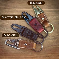 three different types of leather keychains on a wooden surface with the words brass, matte black, nickel and brass