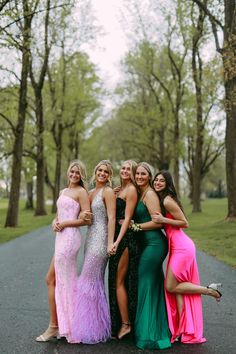 four women in long dresses posing for the camera on a road with trees and grass behind them