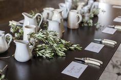 the table is set with white vases and silverware, greenery, and place cards