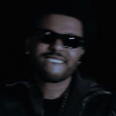 a man wearing sunglasses and smiling at the camera with dark lighting in the back ground