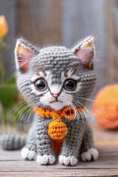 a crocheted kitten wearing a scarf and sitting on a wooden surface with flowers in the background