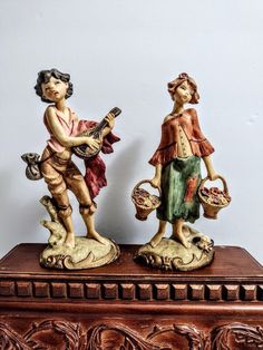 two figurines are standing on top of a wooden box, one holding a basket and the other carrying a bird