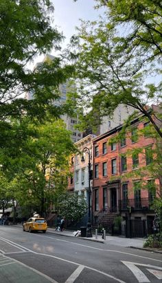 a yellow taxi driving down a street next to tall buildings with lots of green trees