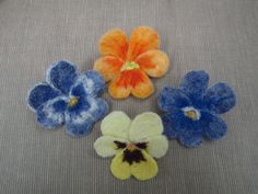 four different colored felt flowers sitting on top of a gray cloth covered surface, with one flower in the center